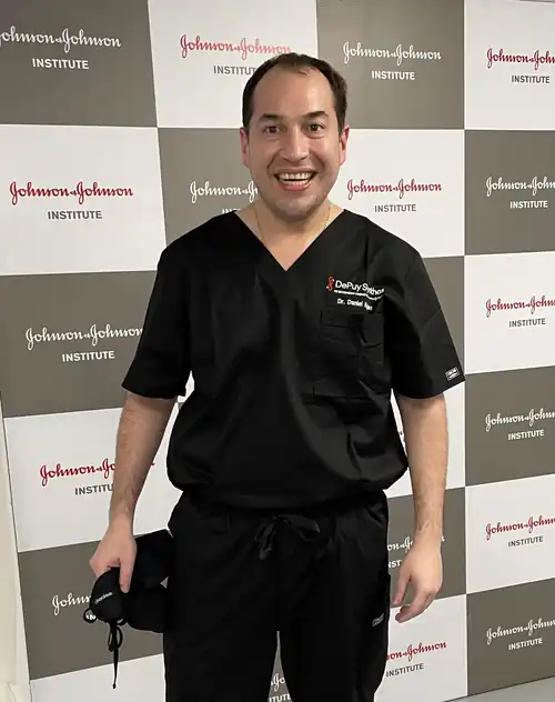 Dr. Daniel Rojas Castillo con uniforme quirúrgico con su nombre, frente al panel de Johnson & Johnson Institute durante el curso cadavérico de 2021
